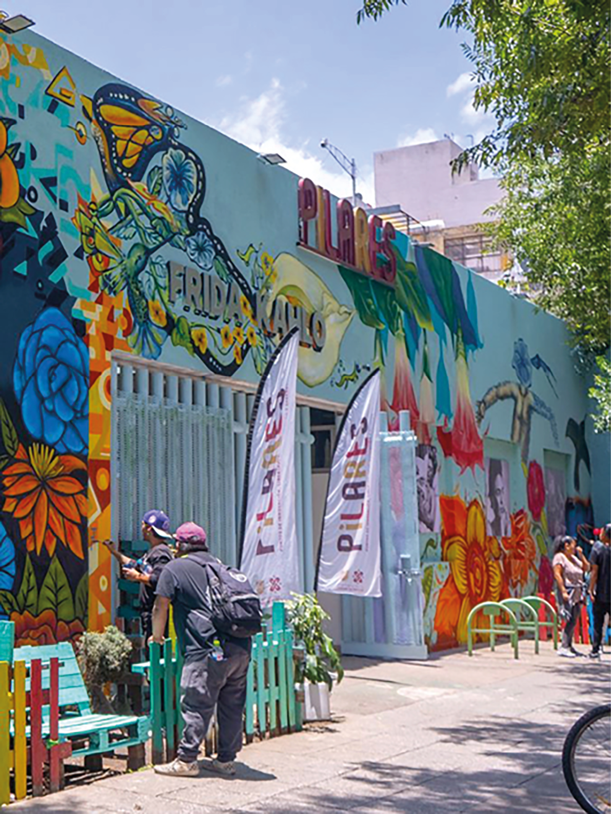 A man is standing outside of a building with a blue bench in front of it. The building has a mural on it and is painted blue and white. There are several people standing outside of the building, and a bicycle is parked nearby. Contenido generado con IA